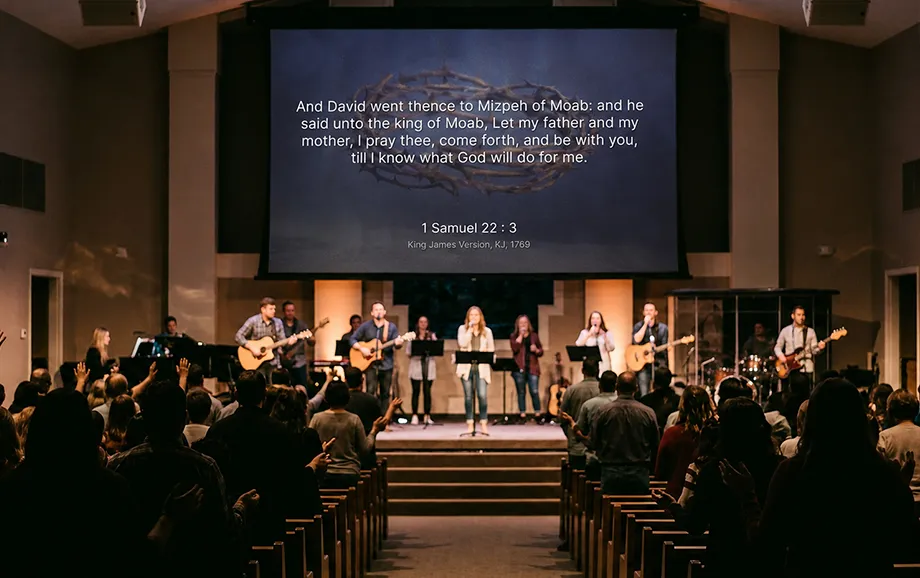Worship band on stage beneath a large screen displaying a Bible passage